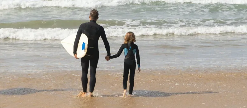 Dad and child wearing wetsuits walking into the sea with a surfboard
